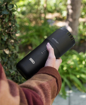 Electric Coffee Travel Mug black V2 - 355ml hålls i hand utomhus med grönskande bakgrund.