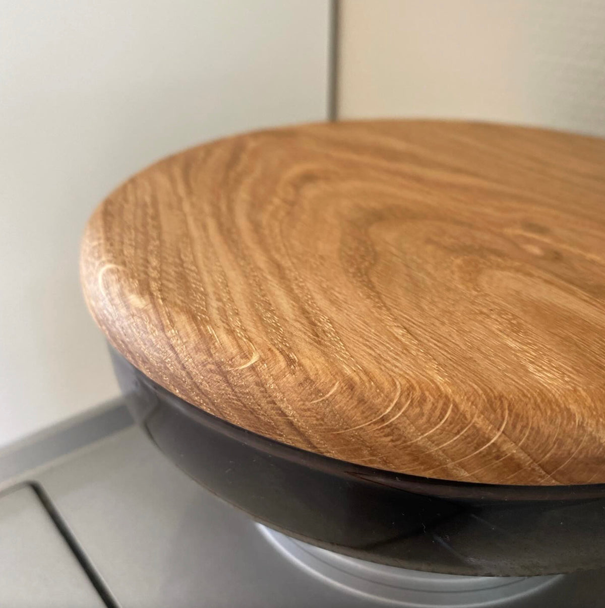 Närbild på ett lock i ekträ med rundad kant vilande på en mörk behållare, mot en ljus bakgrund.