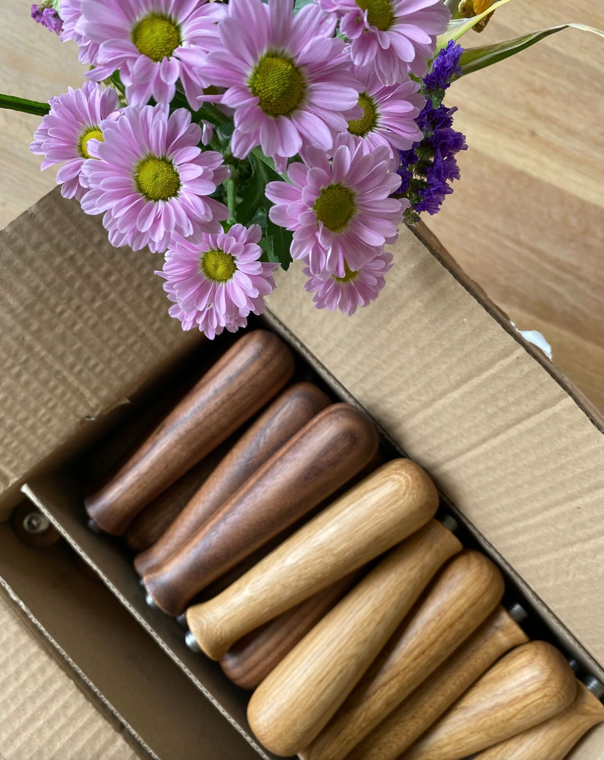 En översikt över trähandtag i en kartong tillsammans med en bukett lila blommor på ett träbord.